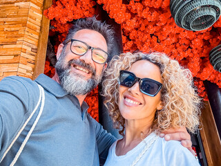 Happy couple middle age taking selfie picture with red flowers in background enjoying tourist vacation outdoor leisure activity in travel lifestyle. Cheerful man and woman sharing content online