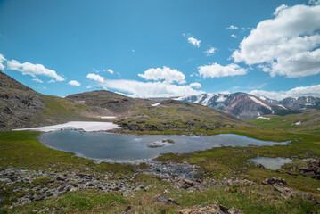 Beautiful small lake with snowfield among green grassy hills and rocks with view to snow-capped mountain range. Scenic landscape with alpine lake and snowy high mountains. Mountain lake in highlands.