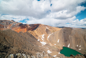 Scenic view from above to green alpine lake in unusual mountains. Colorful landscape with sunlit pass and sharp rocky mountain of vivid colors under cloudy blue sky. Rocks and sheer crags in sunlight.