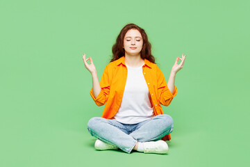 Full body young ginger woman wear orange shirt white t-shirt casual clothes sits hold hands in yoga om aum gesture relax meditate try calm down isolated on plain green background. Lifestyle concept.
