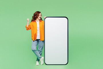 Full body young woman in orange shirt white t-shirt casual clothes big huge blank screen mobile cell phone smartphone with area do winner gesture isolated on plain green background Lifestyle concept
