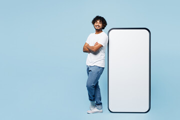 Full body side view young Indian man he wear white t-shirt casual clothes stand near big huge blank screen mobile cell phone smartphone with area isolated on plain blue background. Lifestyle concept.