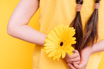 Little girl holding a yellow flower behind her back against a yellow background.
