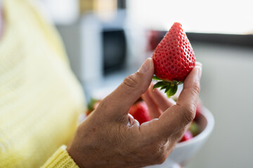 Close-up on senior woman's hand in home kitchen holding a red fresh ripe strawberry. Healthy eating...