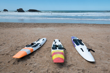 Caiaques na areia em frente para a praia com alguns surfistas e banhistas a aproveitarem as ondas do mar num dia ensolarado - obrazy, fototapety, plakaty