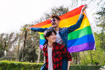 Latino and Chinese gay couple joyfully wave a large LGBT flag, embodying freedom and love.