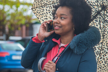 afro woman on the street walking with umbrella using mobile phone