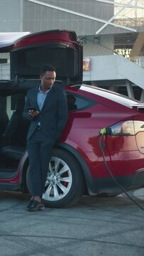Vertical Screen: African man waiting while his electric car charging. Charging car. Full length portrait of successful businessman in suit using modern smartphone for checking working emails while