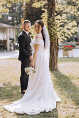 Naklejka premium A bride and groom are posing for a picture in a park. The bride is wearing a white dress and holding a bouquet, while the groom is wearing a suit. The scene is bright and cheerful