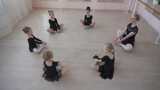 Caucasian woman and five little girls sit in a circle and do stretching at a ballet school. 