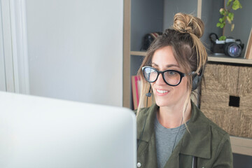 Smiling millennial woman in headphones and glasses sits at desk and talks to customer taking notes, happy young blonde woman works at home answering people for purchase orders using computer