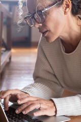 Concentrated blonde woman with glasses working on laptop on the floor of her living room. Middle-aged female typing on computer, technology concept. People browsing the web and chat online. business