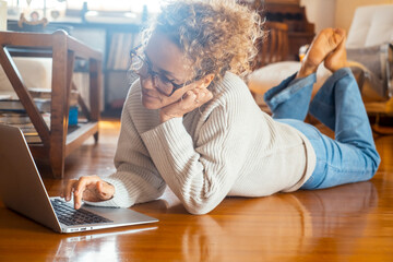 A curly blonde woman with eyeglasses lying on the ground and chooses purchases to make in the online store. Shopping and leisure concept. Relaxed lady working on laptop at home. window in background
