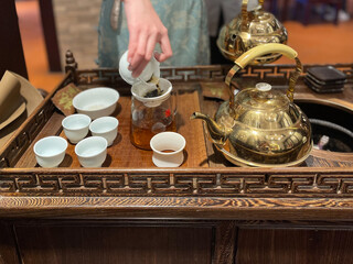 A Chinese lady dressed in traditional Chinese dress serving gourmet tea with a gold color metal pot and Chinese tea cups
