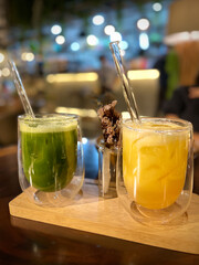A glass of cold orange juice with ice and a glass of healthy green juice with glass stirs at a restaurant setting with blurry background