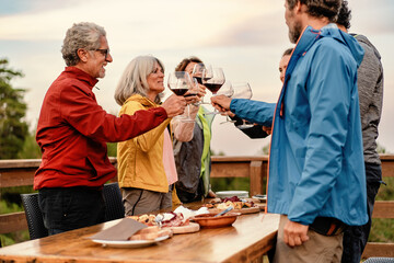 A group of mature friends - toasting with wine glasses at an outdoor table, surrounded by nature at sunset - joyful, festive gathering.