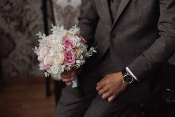 A man in a suit holding a bouquet of flowers. The man is wearing a watch and is sitting on a chair