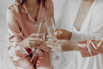 Two women are sitting on a bed, holding champagne glasses and toasting to each other. Scene is celebratory and joyful