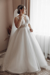 A woman in a wedding dress is standing in front of a window. She is holding a cell phone in her hand
