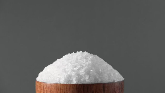 White table salt granules falling into a pile. Wood bowl. Gray color studio background. Condiment particles dropping. Clean cosmetic bath salt pouring footage. Closeup of grained sea salt pieces hill