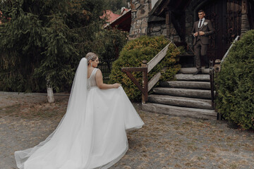 A bride is walking down the stairs in her wedding dress. A groom is waiting for her at the top of the stairs