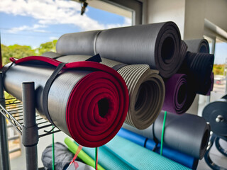 Assorted yoga mats neatly stacked in a rack near the window of a gym.