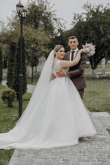 Fototapeta premium A bride and groom are posing for a picture in a park. The bride is wearing a white dress and the groom is wearing a suit. They are holding hands and the bride is holding a bouquet