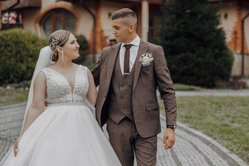 A bride and groom are walking down a path in front of a house. The bride is wearing a white dress and the groom is wearing a suit