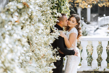 A man and a woman are kissing in front of a white bush. The man is wearing a black shirt and the...