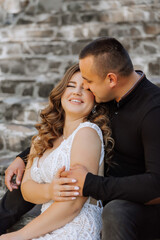 A man and a woman are sitting on a stone wall, kissing each other. The woman is wearing a white dress and the man is wearing a black shirt. Scene is romantic and intimate