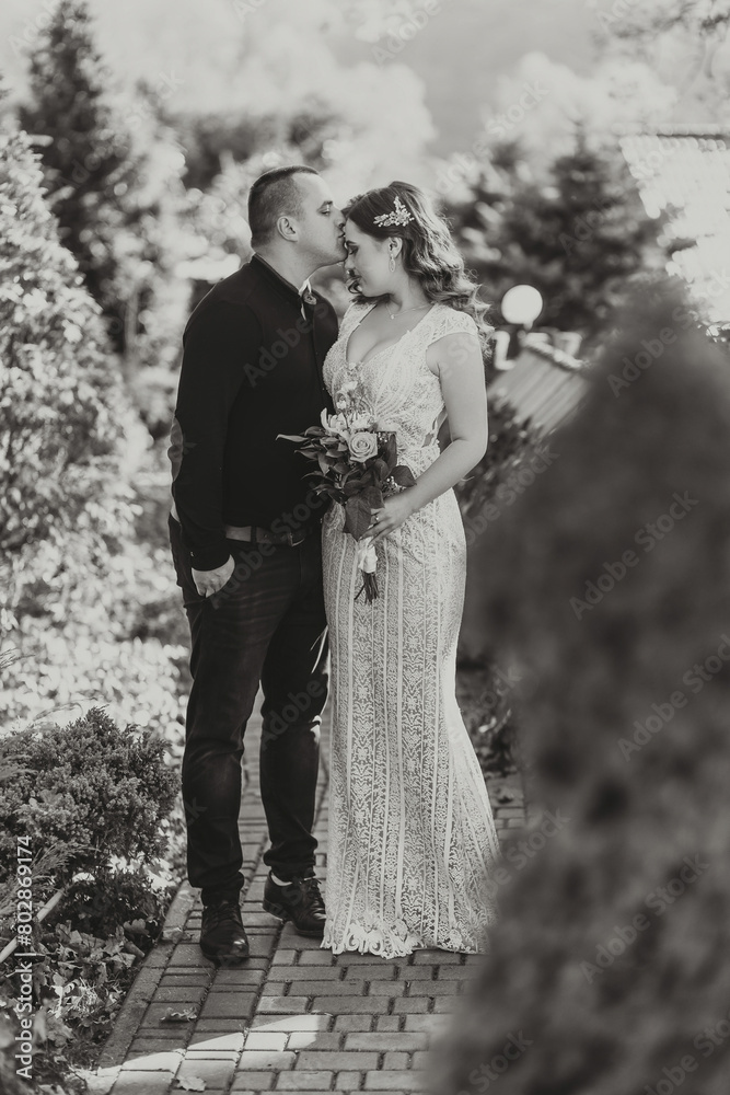 Wall mural A man and woman are kissing in front of a bush. The man is wearing a black shirt and the woman is wearing a white dress