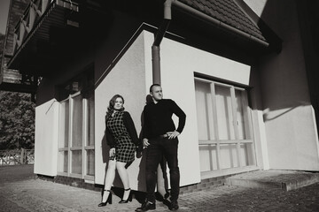 A man and woman stand in front of a building, with the man wearing a black shirt and the woman...