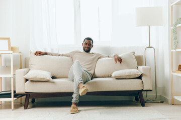 Relaxed African American Man Sitting on Sofa at Home, Lost in Thought A young adult African...