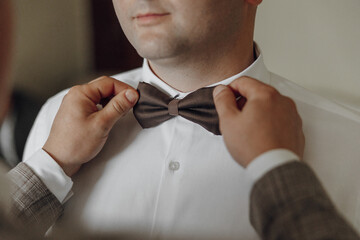 A man is getting ready to wear a bow tie. A man is helping him tie it. The man is wearing a white...