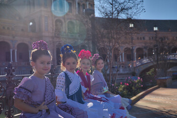 Four girls dancing flamenco, looking at camera, in typical flamenco costumes sitting on a bench in a nice square in Seville, receiving sun rays. Dance concept, flamenco, typical Spanish, Spain.