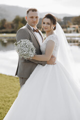 A bride and groom are posing for a picture in front of a lake. The bride is wearing a white dress...
