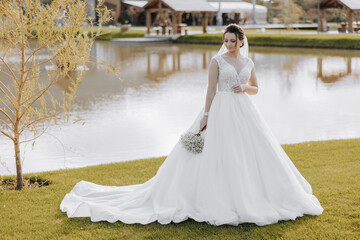 A woman in a white wedding dress stands in front of a lake. She is holding a bouquet of flowers