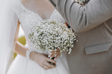 A bride and groom are holding hands and the bride is holding a bouquet of white flowers. Scene is...