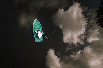 Valmiera, Latvia - August 19, 2023 -  a bride and groom sitting together in a green rowboat on a calm, reflective water body, with trees and clouds mirrored in the water.