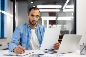 Fototapeta premium Portrait of serious hispanic man working in office with laptop and documents, writing down data and information. Looks confidently into the camera