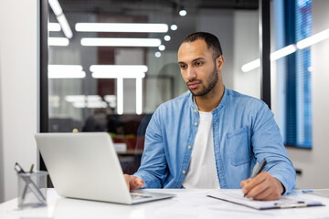 Fototapeta premium Serious young hispanic man working in office at desk with laptop and making notes in notebook, watching online training and meeting
