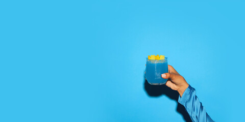 Female hand holding sweet and sour blue tropical cocktail with pineapple slice against bright blue background. Concept of alcohol and non-alcohol drink, party, holidays, bar. Poster. Copy space for ad - obrazy, fototapety, plakaty