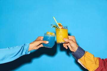 Male and female hand clinking glasses with blue and yellow sweet and sour cocktail against blue background. Cheers. Concept of alcohol and non-alcohol drink, party, holidays. Poster. Copy space for ad - obrazy, fototapety, plakaty