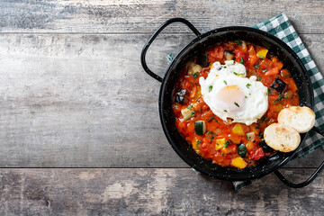Vegetable pisto manchego with tomatoes, zucchini, peppers, onions,eggplant and egg, served in frying pan on wooden table. Top view. Copy space