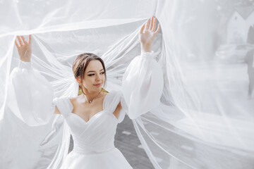 A woman in a white dress is standing in front of a white curtain. She is holding the curtain open,...