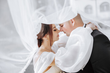 A bride and groom are embracing each other under a white veil. The bride is wearing a necklace and...