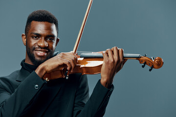 African American male musician in elegant black suit playing the violin on gray background,...