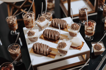 A table full of desserts, including cakes and pastries, is set up for a party. The desserts are arranged in small cups and bowls, and there are spoons available for guests to use