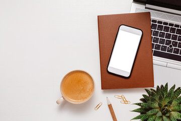 Home office workplace. Smartphone with white screen on notepad, coffee, plant, laptop and office supplies. Cozy home office, smartphone for promotion, web site and design
