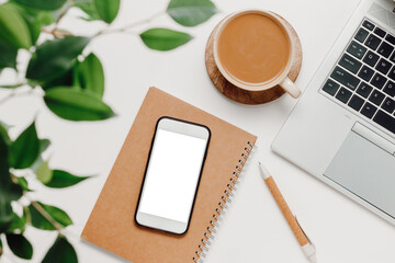 Smartphone with white screen on notepad, coffee, laptop and green leaves. Aesthetic home office, smartphone for promotion, web site and design